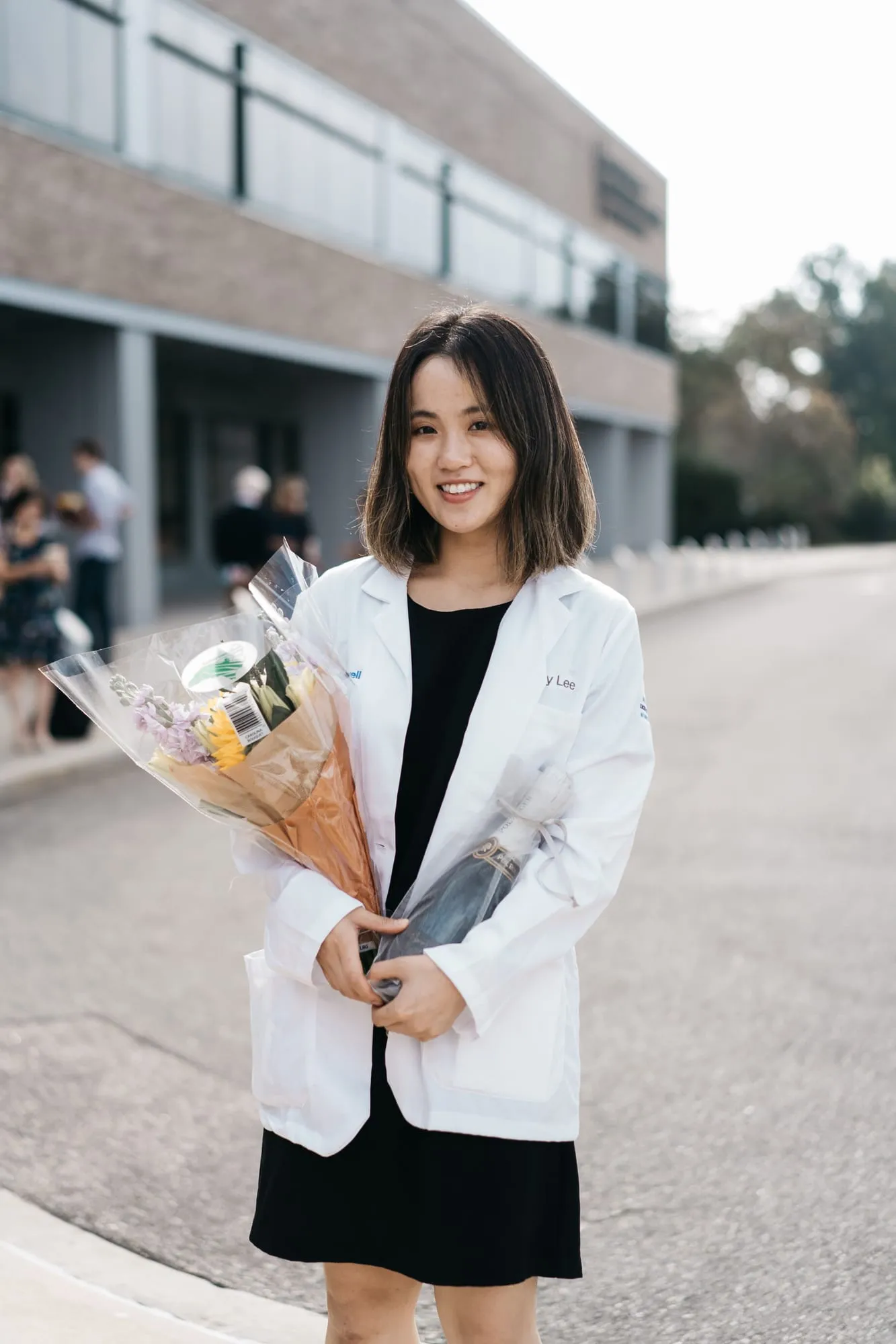 A graduate in a white coat holding a wrapped bouquet outside a campus building.