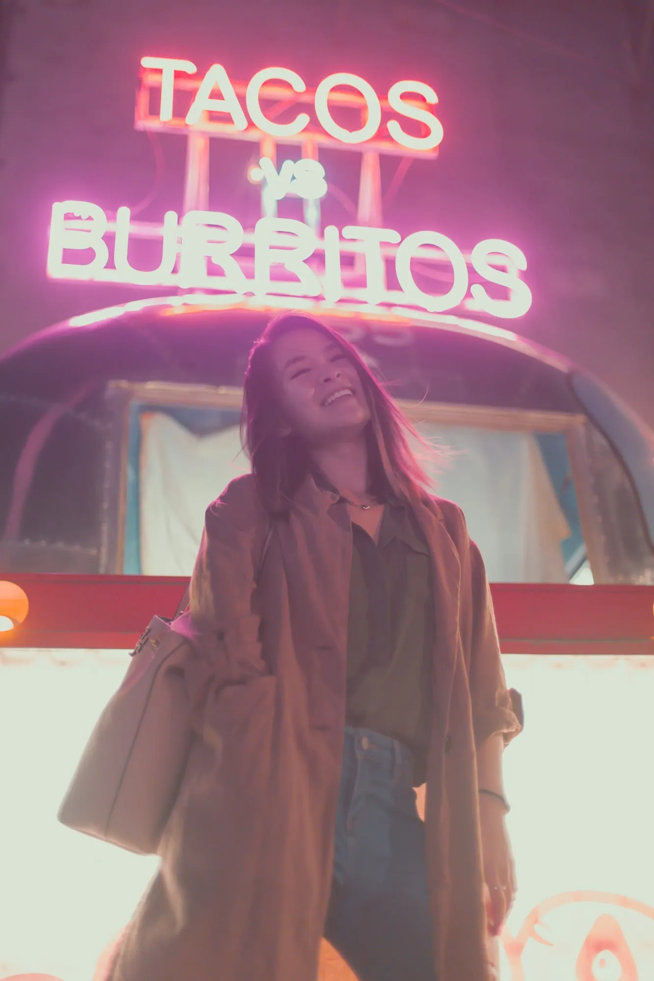 A woman laughing in front of a pink neon 'Burritos' sign on a food truck at night.