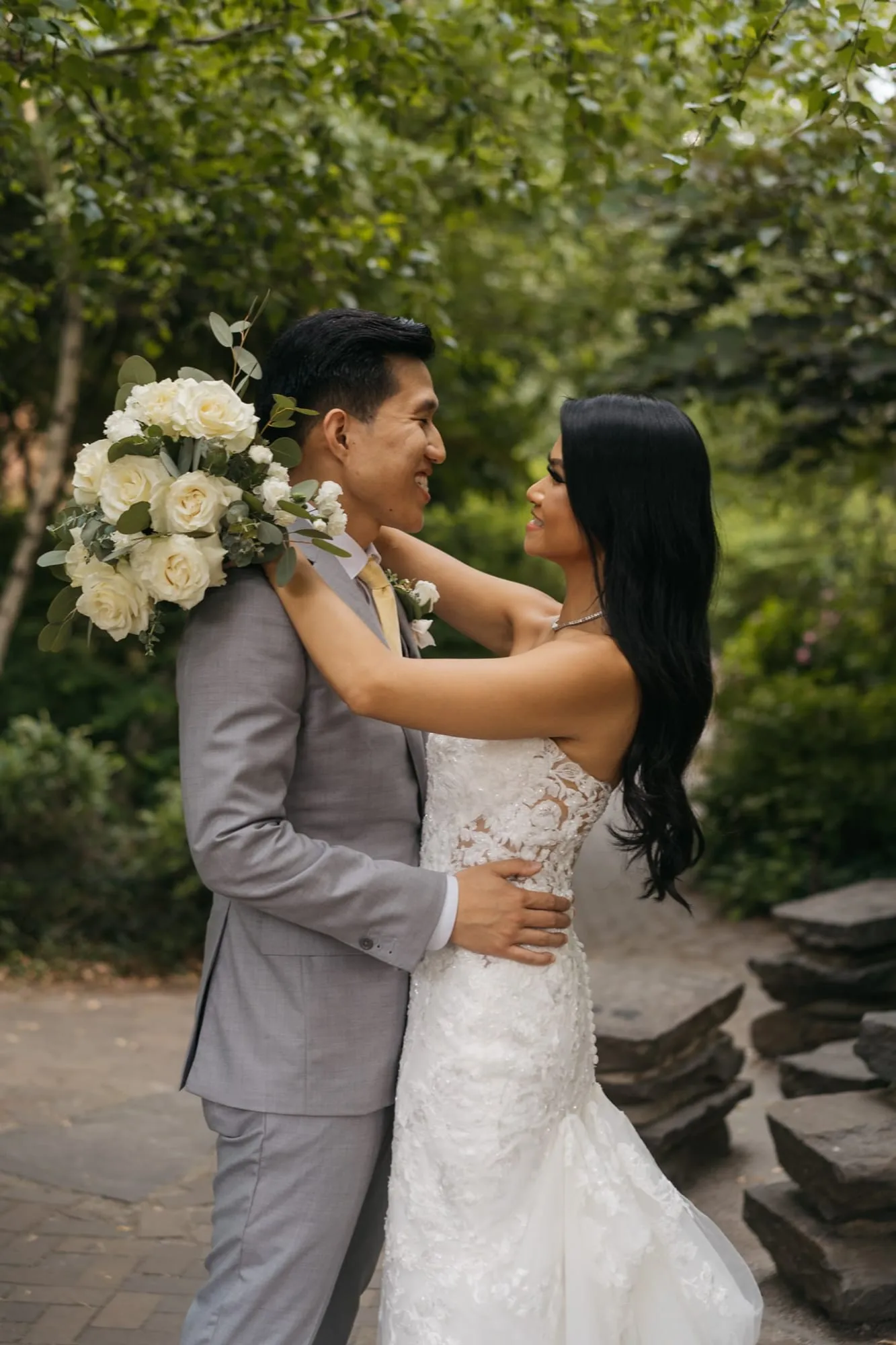 A bride holding a white bouquet and a groom in a grey suit embracing on a garden path.